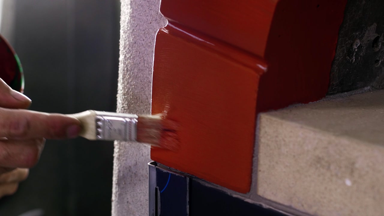 Close-up of hand brushing glossy terracotta paint onto curved edge of pizza oven structure.