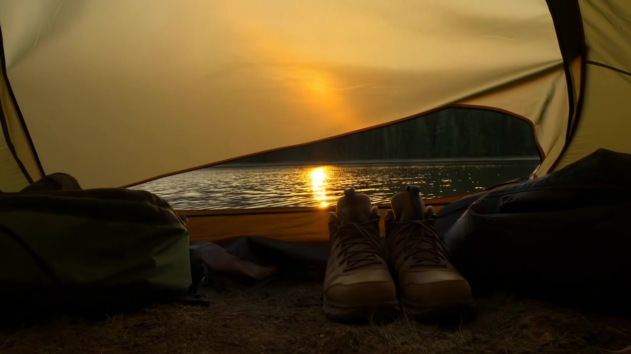 Lavt perspektiv indefra teltet viser snørede vandrestøvler og campingudstyr nær indgangen, mens varmt solnedgangslys sænker sig mod horisonten og maler den rolige sø med bløde refleksioner.