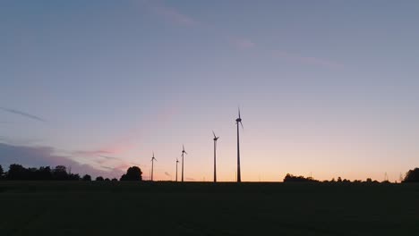 Aerial-establishing-view-wind-turbines-generating-renewable-energy-in-a-wind-farm,-evening-after-the-sunset-golden-hour,-countryside-landscape,-high-contrast-silhouettes,-drone-shot-moving-backward