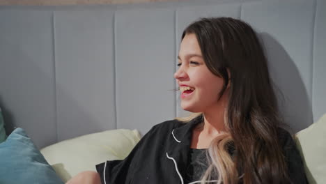 Close up of cheerful young woman with long dark hair laughing while eating snack and watching movie, she sits comfortably on bed surrounded by soft pillows and blankets in relaxed bedroom setting