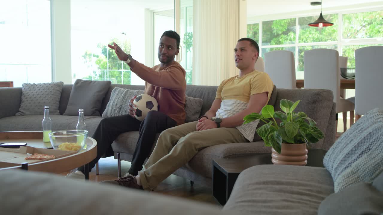 Diverse mannelijke vrienden thuis die een bal vasthouden, naar een voetbalwedstrijd kijken, wijzen en juichen met snacks.