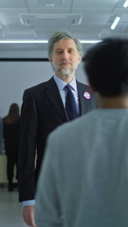 Portrait-of-businessman,-United-States-of-America-elections-voter.-Mature-man-stands-in-a-modern-polling-station,-poses-and-looks-at-camera,-smiles.-Background-with-voting-booths.-Civic-duty-concept.