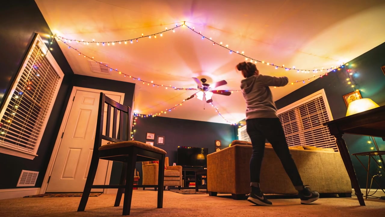 Using wooden chair woman in hoodie hanging multicolor string lights to living room ceiling for mood