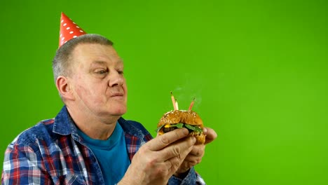 senior adult male caucasian ethnicity in birthday cap blows out candles on hamburger instead of birthday cake and eats burger with delight.
