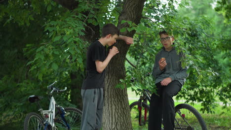 two young siblings enjoy a break while eating snacks under a tree in a peaceful forest, one sibling leans on the tree while the other sits on his bicycle, both surrounded by vibrant greenery