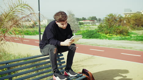 man writing notes with a pen while studying sitting on a bench outdoor