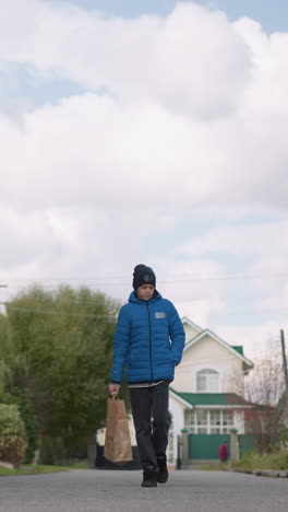 niño en chaqueta azul paseando pensativo a lo largo de una calle residencial llevando una bolsa de compras, el fondo cuenta con árboles de otoño, casas suburbanas, postes de servicios públicos, y alguien caminando en la distancia