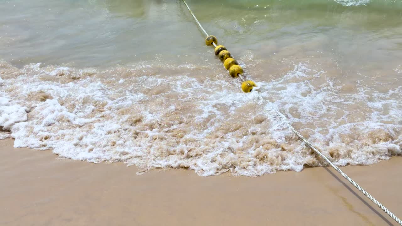 Ocean waves wash over a yellow buoy rope line on a sandy tropical beach shore