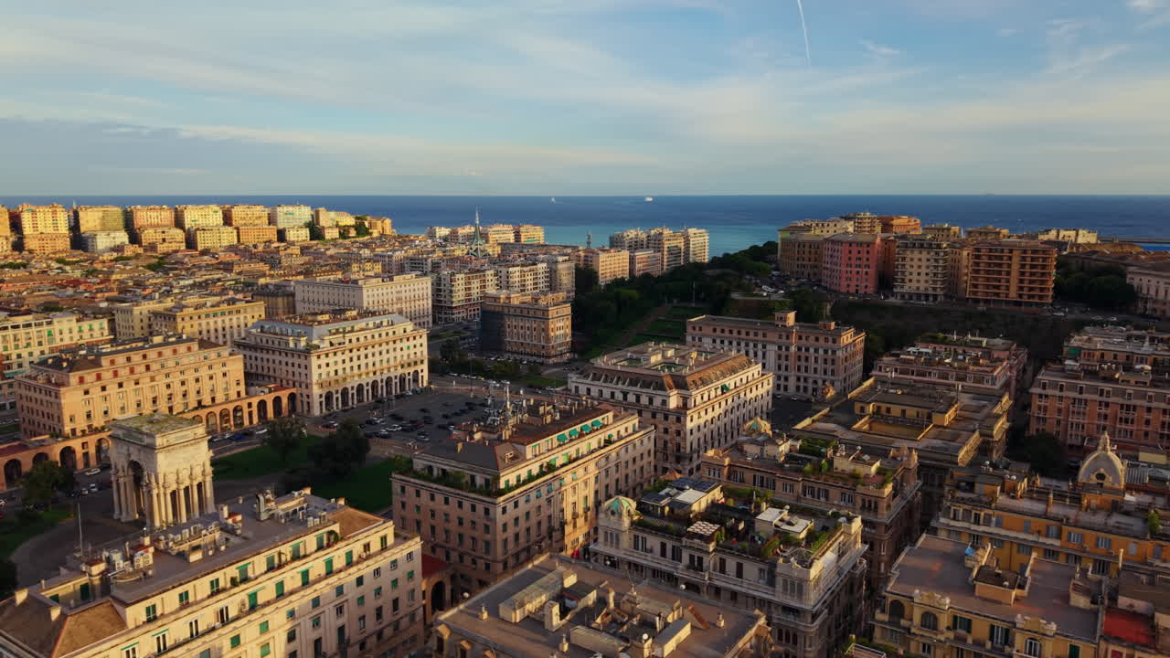 Drone makes a lateral move above Genoa city center at sunset, showing colorful symmetrical buildings, the sea and the sky glowing in the background