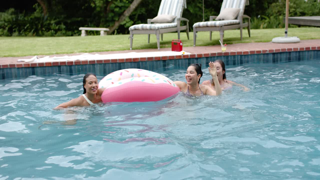 Diverse women reaching for and pushing pink donut in backyard pool, adjusting shades, steadying it