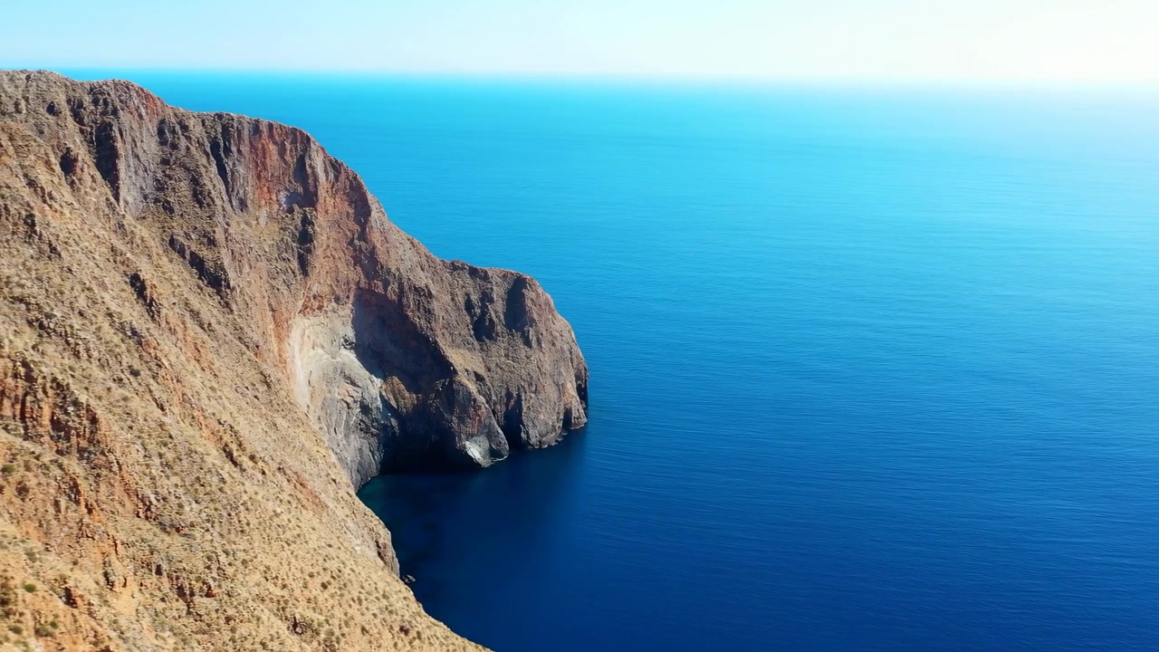 Scogliera remota che si affaccia su un mare blu profondo, ripidi pendii brulli che scendono nell'oceano aperto, orizzonte ampio.