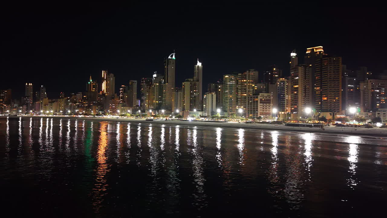 Smooth right-tracking drone shot captures Camboriú’s skyscrapers casting shimmering reflections on night waters, merging urban grandeur with coastal calm. Ideal for luxury travel or city branding