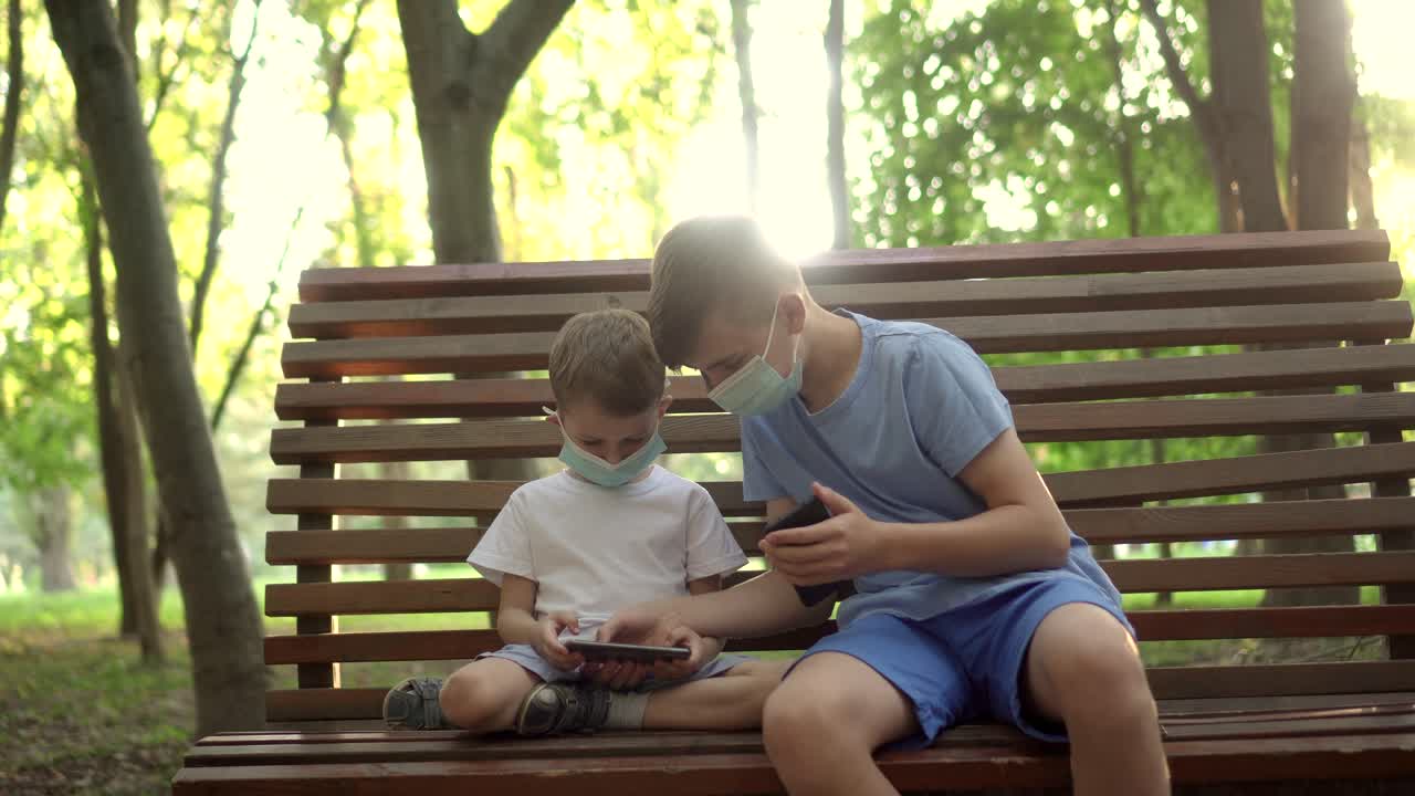 niños con máscaras médicas juegan con teléfonos inteligentes en el parque 02