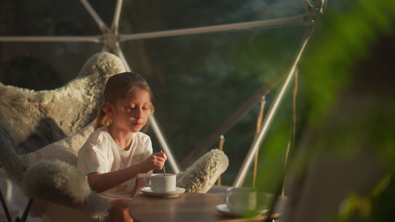Relaxed child stirs drink with spoon at glamping. Quiet girl has leisurely breakfast sitting at table in transparent tent during holiday slow motion