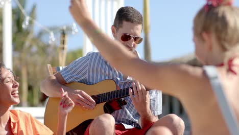 Fröhliche,-Vielfältige-Gruppe-Von-Freunden,-Die-Am-Strand-Mit-Palmen-Und-Strandhaus-Sitzen-Und-Gitarre-Spielen