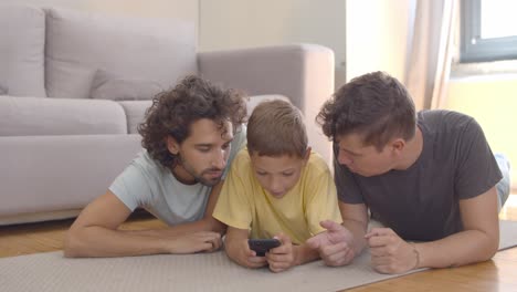 Focused-boy-lying-on-floor-near-dads-and-using-app-on-cell