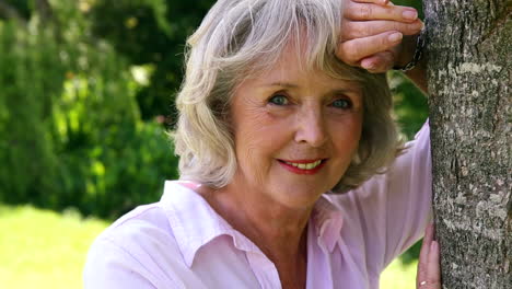 retired woman leaning against tree smiling at camera