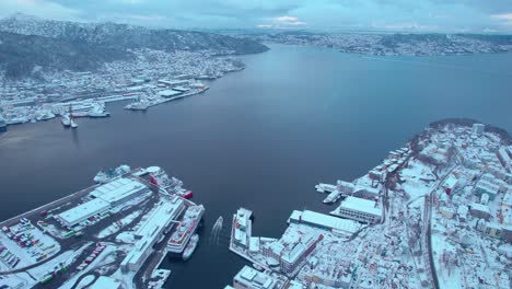 Boat-exits-harbor-leaving-Bergen-Norway-covered-in-snow,-panoramic-aerial