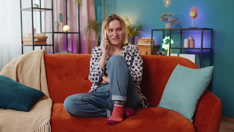 Smiling-young-Caucasian-woman-making-mobile-phone-conversation-with-friends-sitting-on-couch-at-home