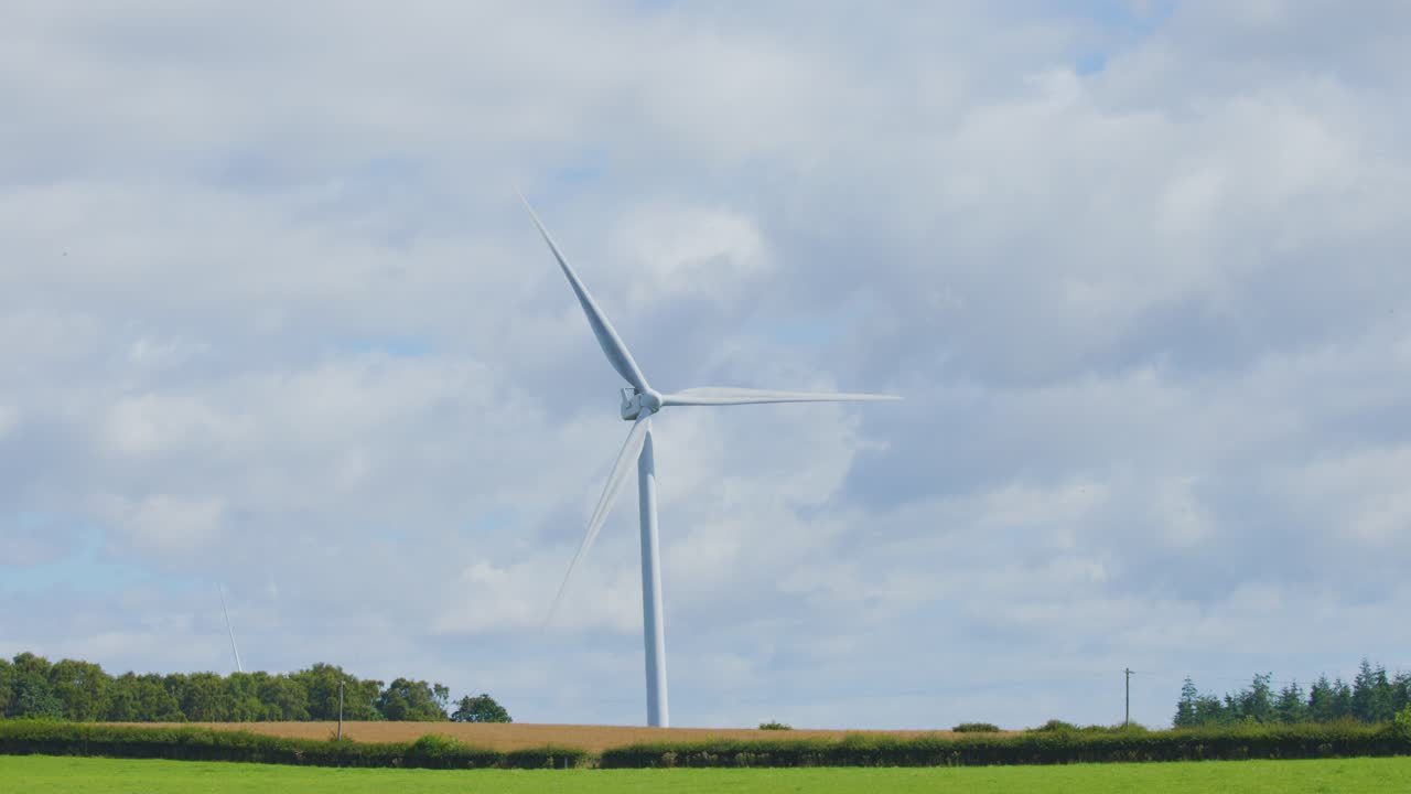 A single wind turbine spins steadily in a rural field under partly cloudy skies, captured in a static wide shot with natural daylight