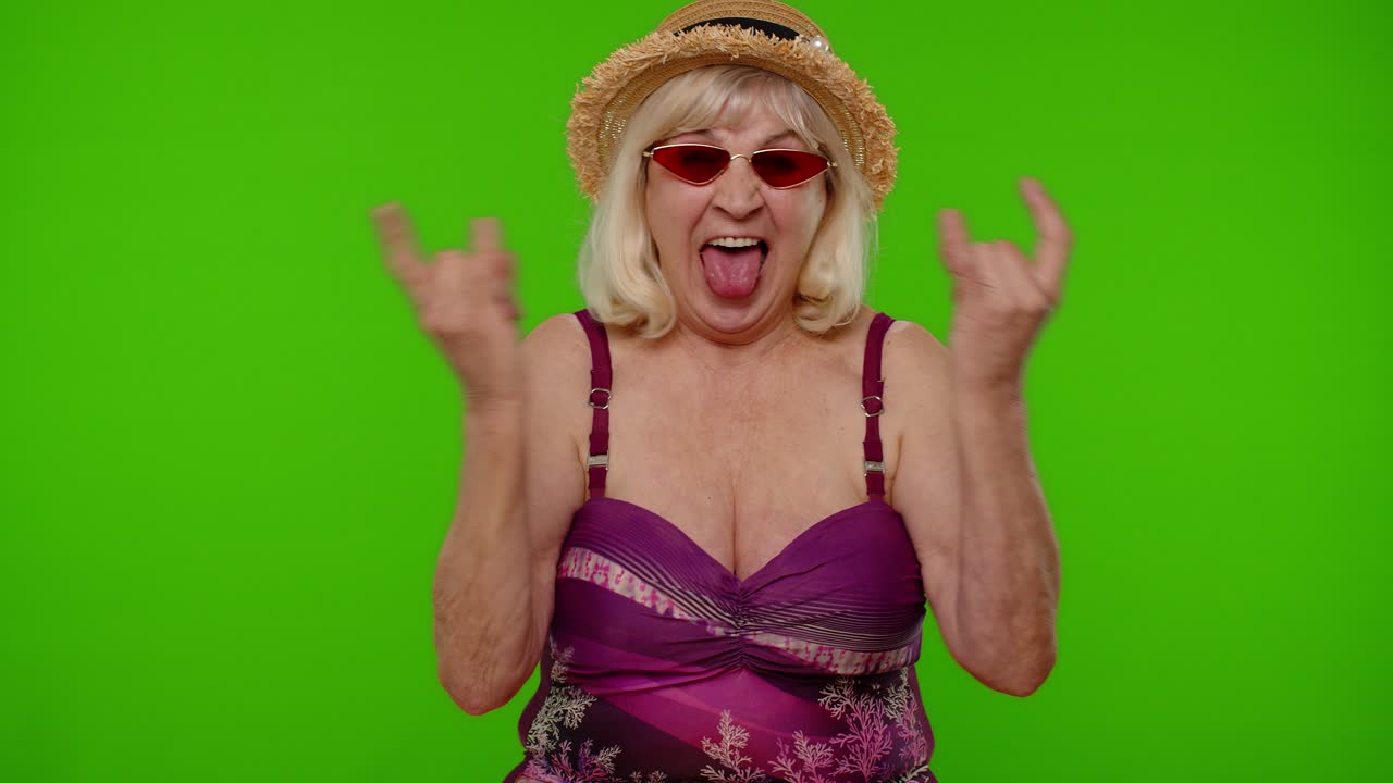 Anziana turista in costume da bagno che balla, festeggia, mostra la lingua e fa il gesto rock and roll con la mano.