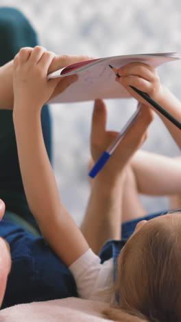cu, concept of distance learning: little girl with her dad lying on floor, shaking her feet, draws in drawing block by pencil, father helps her, sunlight, in footside and jeans, do homework closeup