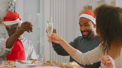 Familia-Multigeneracional-Con-Gorros-De-Papá-Noel-Haciendo-Un-Brindis-Durante-La-Comida-Navideña-En-Casa-Juntos