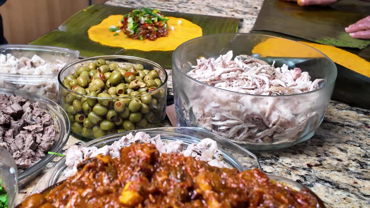 Nærbillede af traditionel samling af venezuelanske hallacas, rigt krydret kødgryderet sammen med skåle med oliven, strimlet kylling, løg, majsdej spredt ud over bananblade, venezuelansk ferieret
