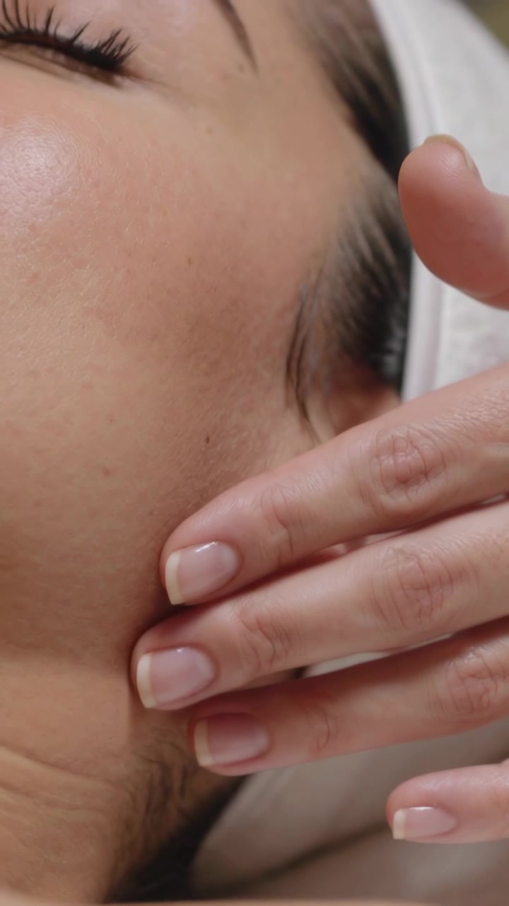 Video verticale: Mano del terapista che tocca e scivola sulla guancia destra in una spa, con una fascia bianca per il drenaggio.