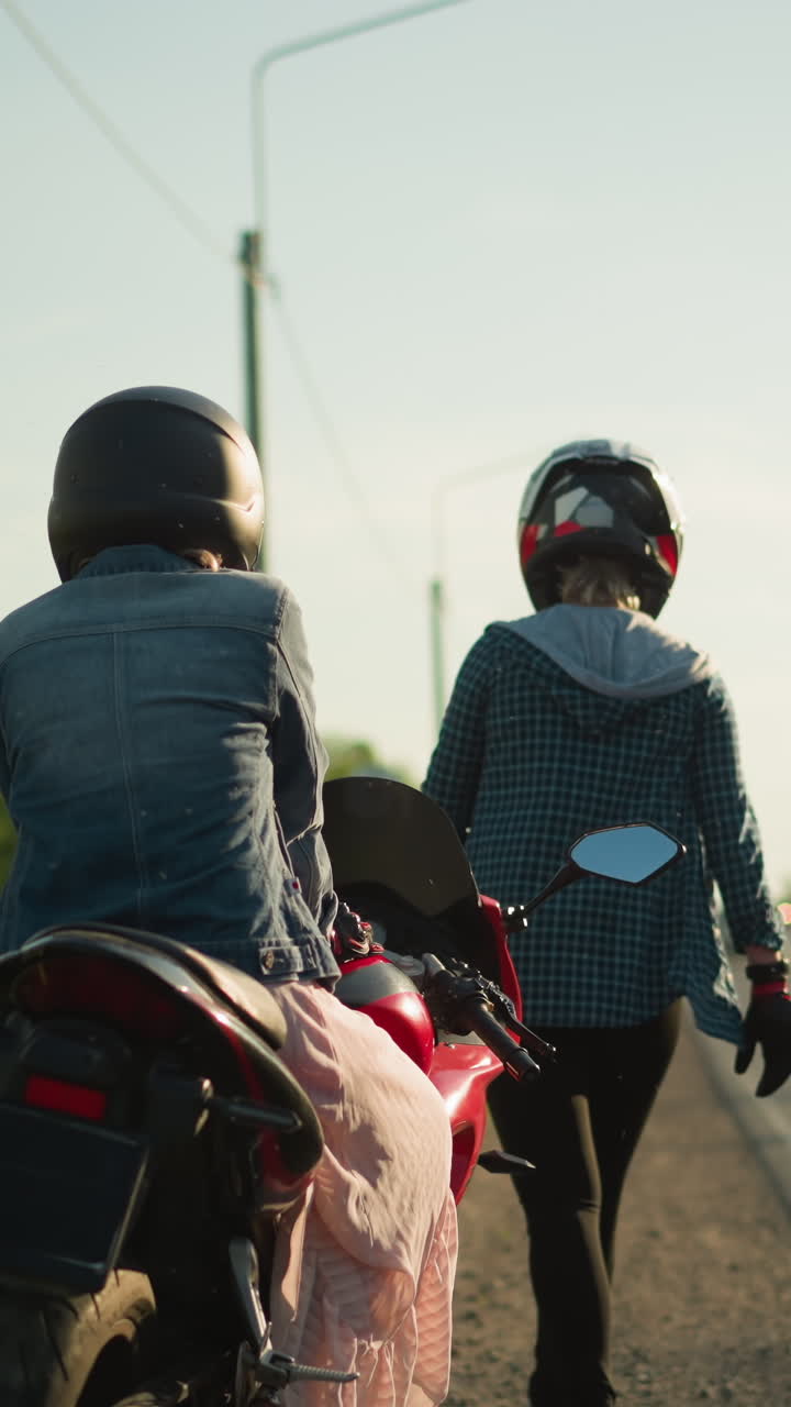 두 명의 헬을 입은 여성이 도로 에 서 있으며, 한 명은 오토바이에 앉아 있고 다른 한 명은 멀리 고 있으며, 배경에는 차가 조용한 도로에서 멀리서 접근합니다.