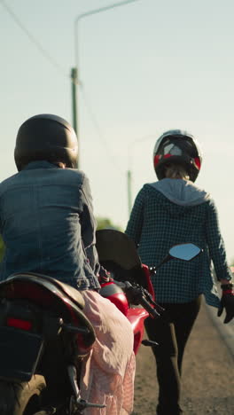 dos mujeres con cascos de pie junto a la carretera con uno sentado en una motocicleta mientras el otro se aleja, en el fondo, los coches se acercan desde la distancia en una carretera tranquila