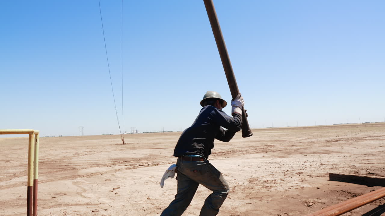A metal tower for drilling oil or gas in the desert. Low angle view. Worker grabs the pipe hanging from rig and carries it aside.