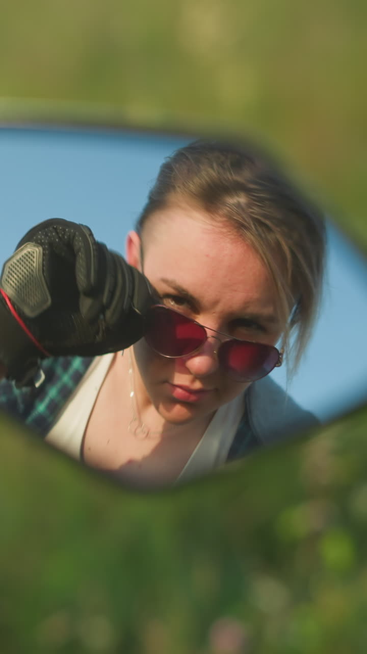 un reflejo en el espejo de una motocicleta que muestra a una mujer con una cálida sonrisa ajustando sus gafas de sol, está en un campo cubierto de hierba