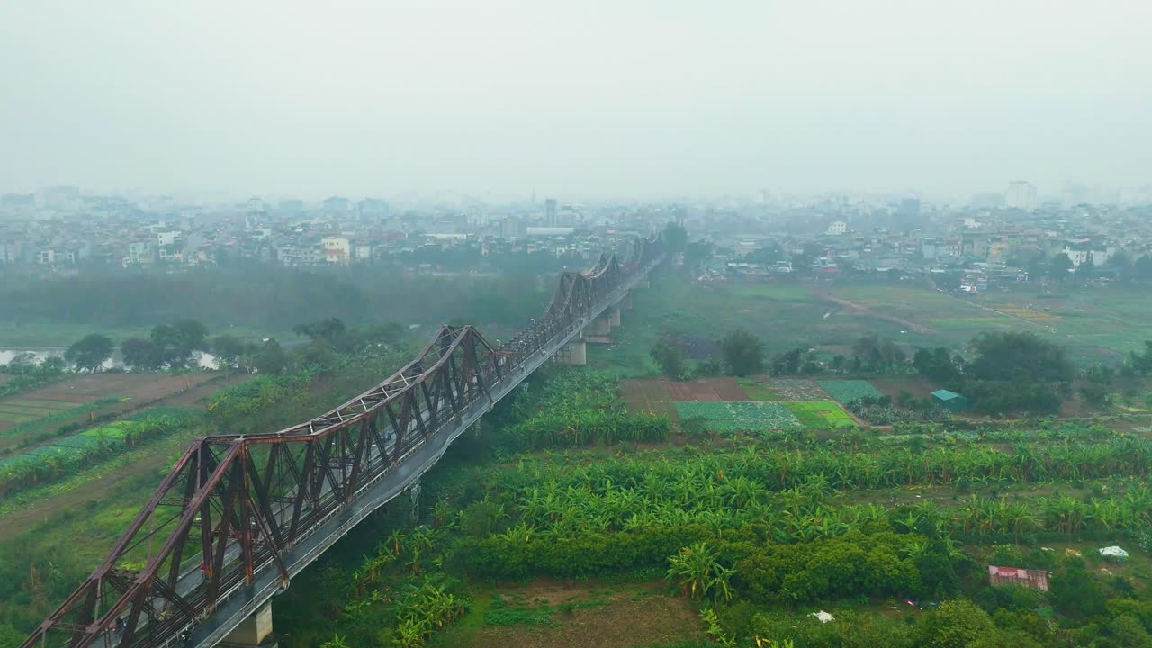 Aerial view of Hanoi's Long Bien Bridge in Vietnam, with a hazy atmosphere caused by extreme pollution. The shot captures the iconic bridge stretching over the green landscape and urban sprawl.