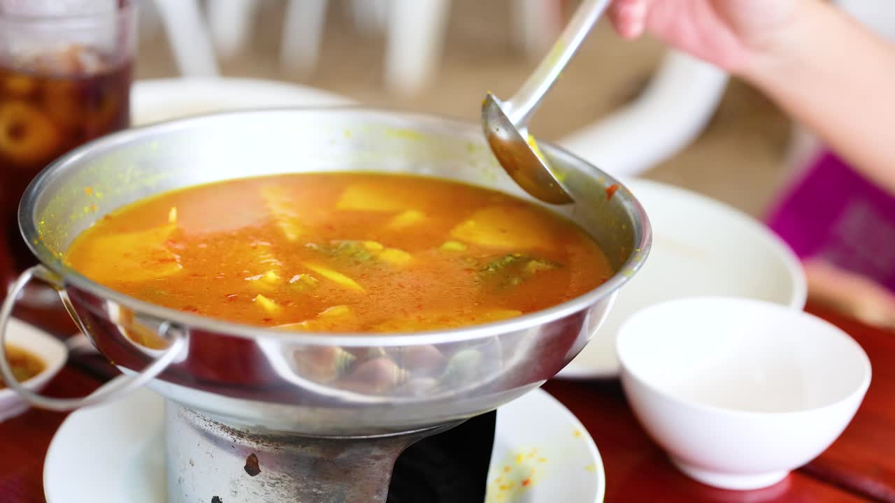 una persona sirve sopa tailandesa caliente de una olla de metal en un entorno de krabi vibrante, mostrando la cultura culinaria local