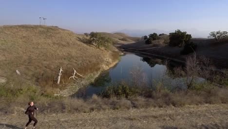 Woman-Running-Alongside-Beautiful-Lake-During-The-Evening-Aerial-Drone