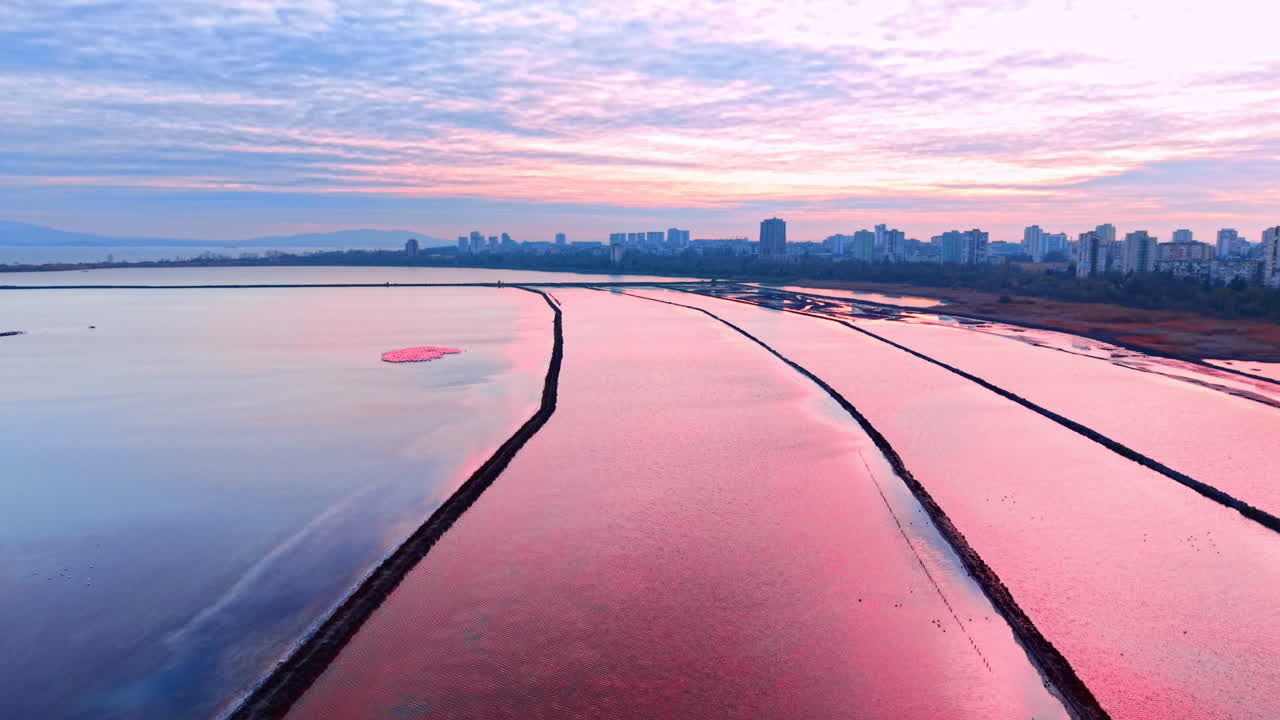 Tanques retangulares de evaporação de sal rosa divididos por caminhos com o horizonte da cidade e o céu do pôr do sol. Vista aérea de salinas rosa em Burgas.