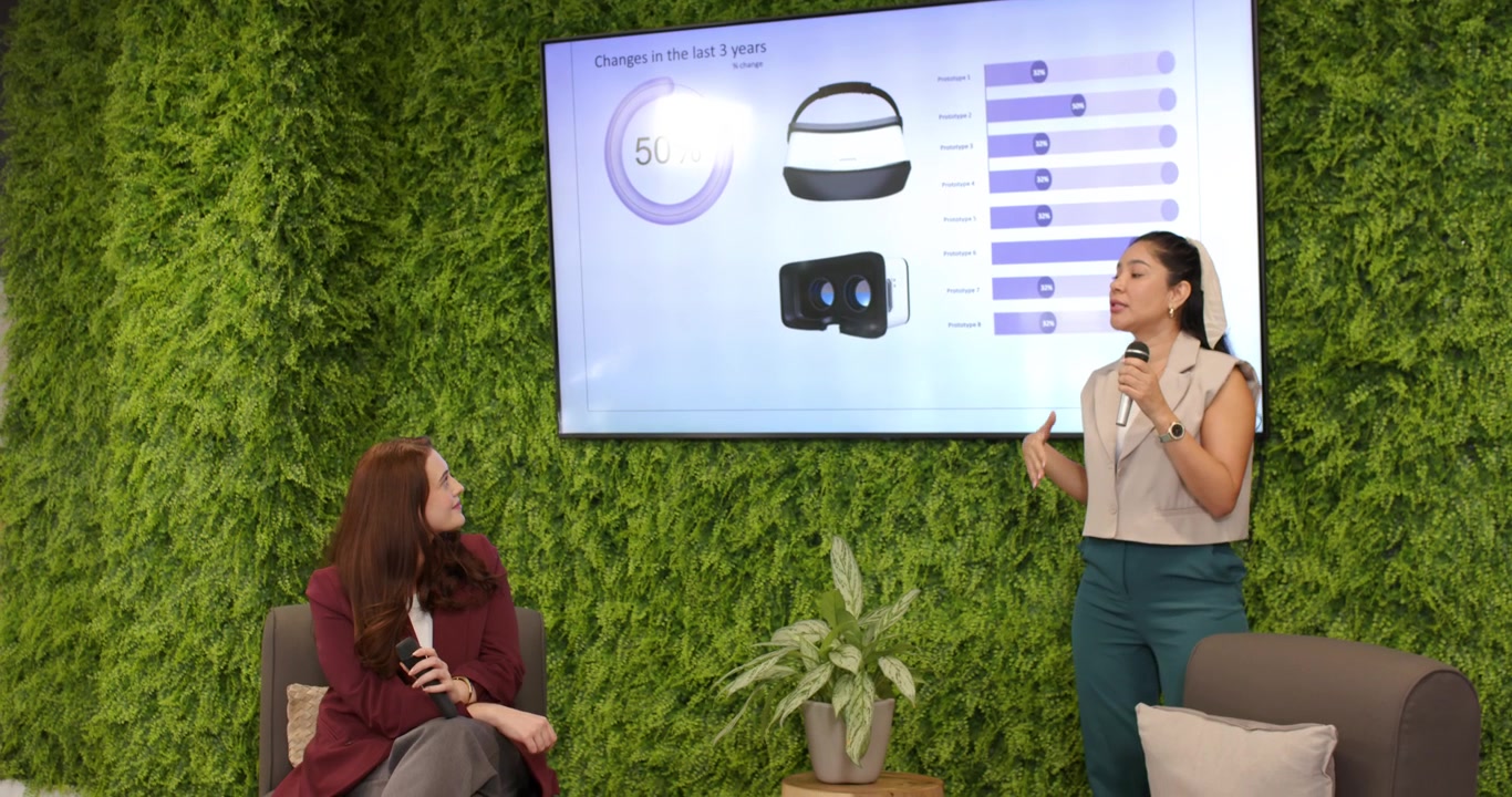 Two women in office host holding mic pointing to slide charts explaining headset data, copy space