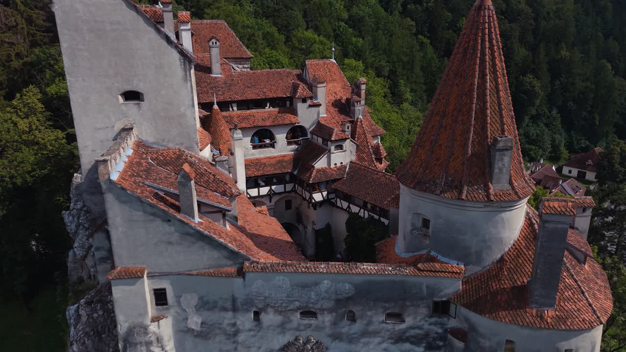 Ripresa con drone che si allontana e si eleva sopra il cortile del Castello di Bran, rivelando tetti di tegole rosse, torri e la foresta circostante