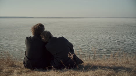 rear view of romantic couple sitting on shore of frozen lake