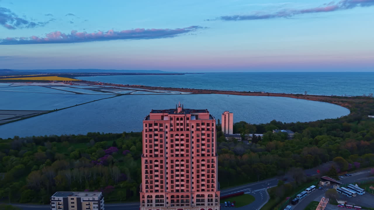 Vista de um edifício alto próximo à água durante o pôr do sol, com céus azuis e colinas distantes ao fundo