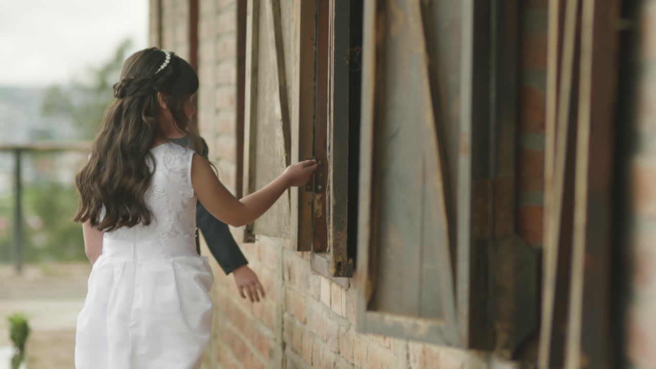 A boy in a suit and a girl in a white dress walk together along a path near the windows of a stable.