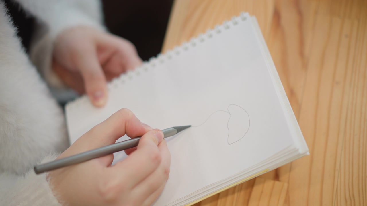 Over-shoulder view of hands drawing light circular shapes on spiral-bound notepad while left hand steadies page on wooden tabletop, with soft indoor light, slight pen blur, and creative focus