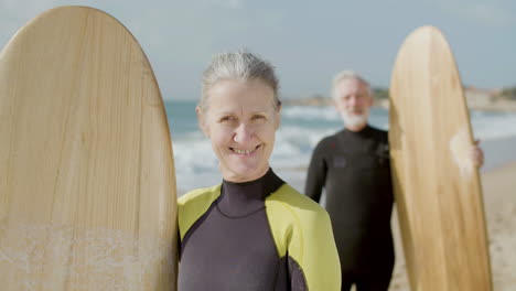 front view of a happy senior woman in wetsuit with surfboard standing on the sandy beach and looking at the camera while her husband standing behind in the blurred background