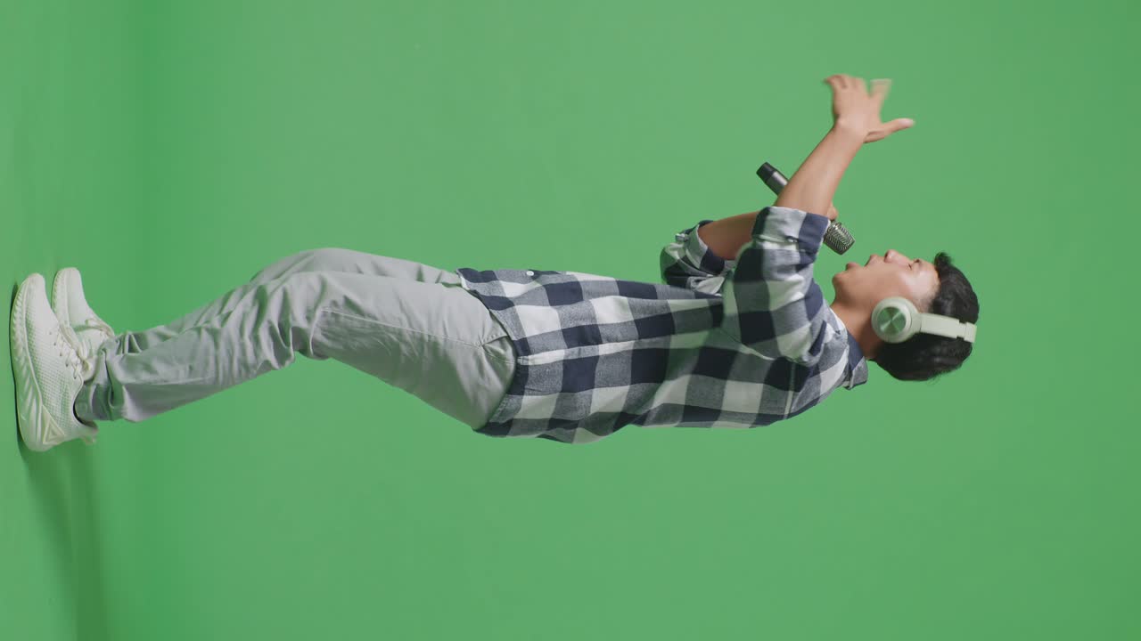 헤드폰을 착용하고 마이크를 들고 녹색 화면 배경에서 하는 젊은 아시아 십대 소년의 전체 몸 측면 시각