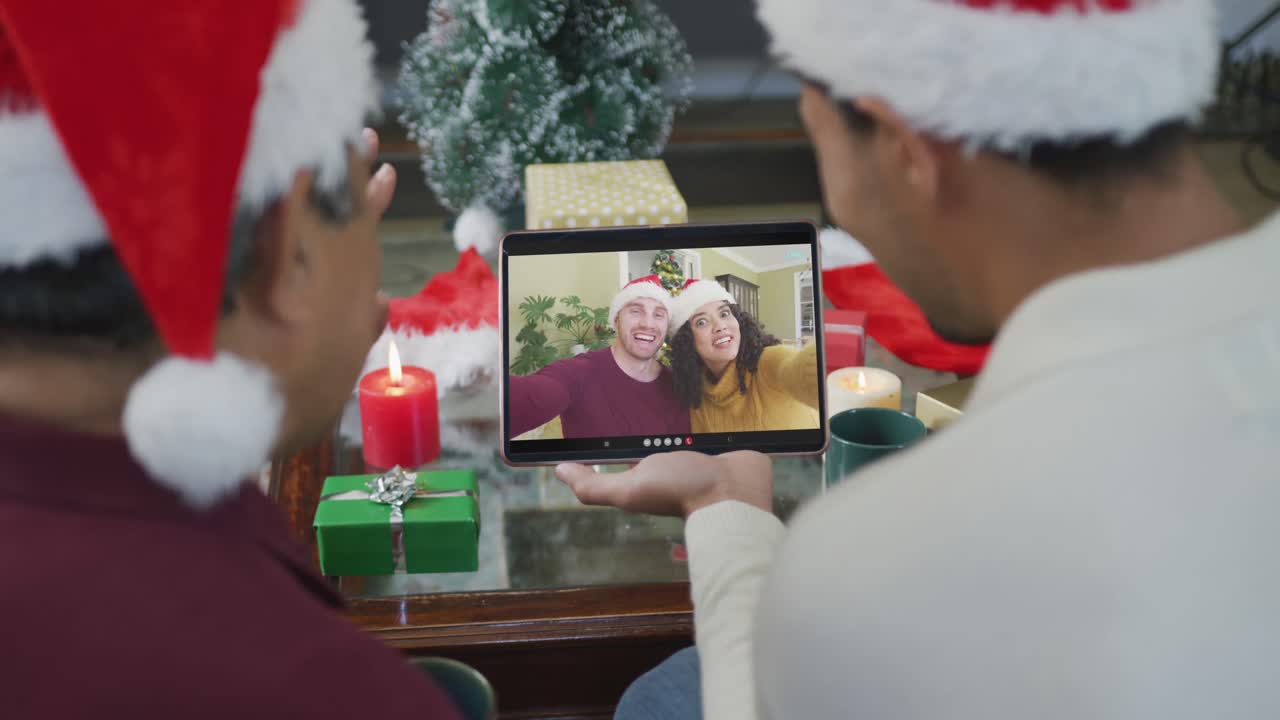padre y hijo biraciales con sombreros de santa usando una tableta para una videollamada de navidad con la pareja en la pantalla