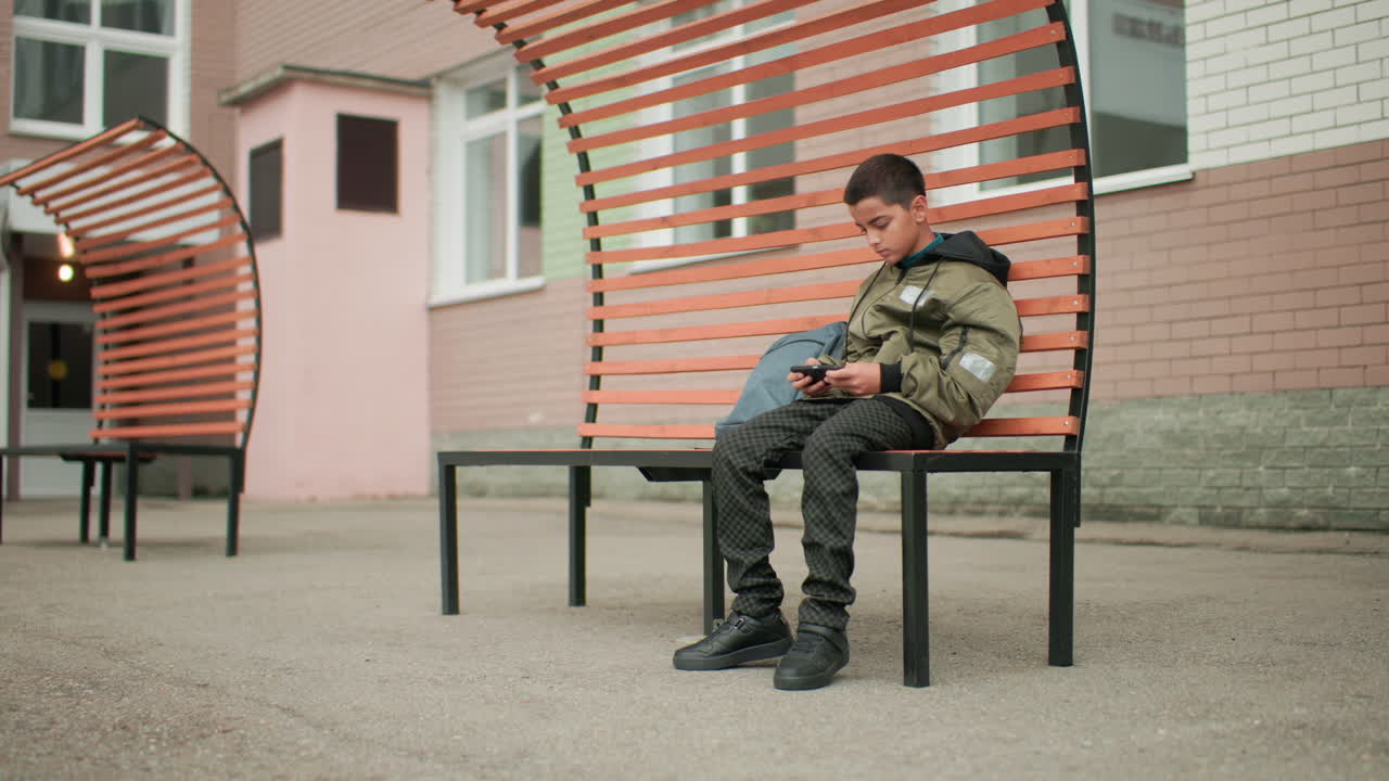 Estudiante sentado en un banco moderno durante el recreo, mirando el móvil concentrado en un juego, con la mochila a un lado cerca del patio del colegio, creando una atmósfera tranquila al aire libre con una postura relajada y energía juvenil