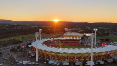drone captures a sweeping aerial view of a brightly lit sports stadium at sunset, with surrounding roads, river, and hinterland in gold coast, australia
