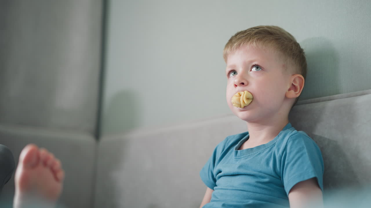 Cute little boy in blue top sitting on bed forcing snack into mouth while gazing thoughtfully at distance, with visible foot of another person nearby