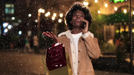 close-up view of african american woman holding shopping bag and talking on smartphone on the street while it‚äôs snowing in christmas